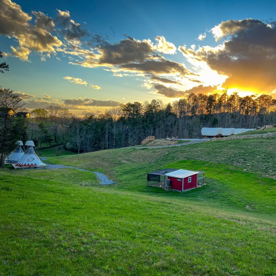 Tipis, wagons, chicken coop, and barn under a beautiful sunset